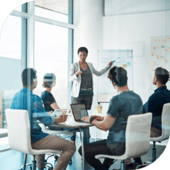 Five colleagues sit around a small conference table in a windowed office focused on a whiteboard presentation.