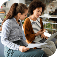 Two women sit side-by-side, one holding a piece of paper and the other using a laptop, and are engaged in a conversation.