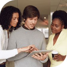 two women stand on either side of a man holding a tablet. The man and the woman to the left of him point at the screen. 